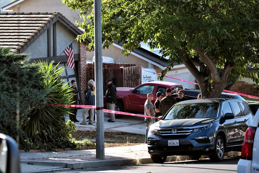 Police tape surrounds a suburban house, with two groups of investigators standing and talking in front of it