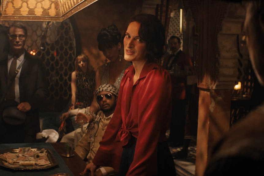 A white woman with short brown hair wears a red silk blouse and commands attention in a roomful of men around a table of cash.