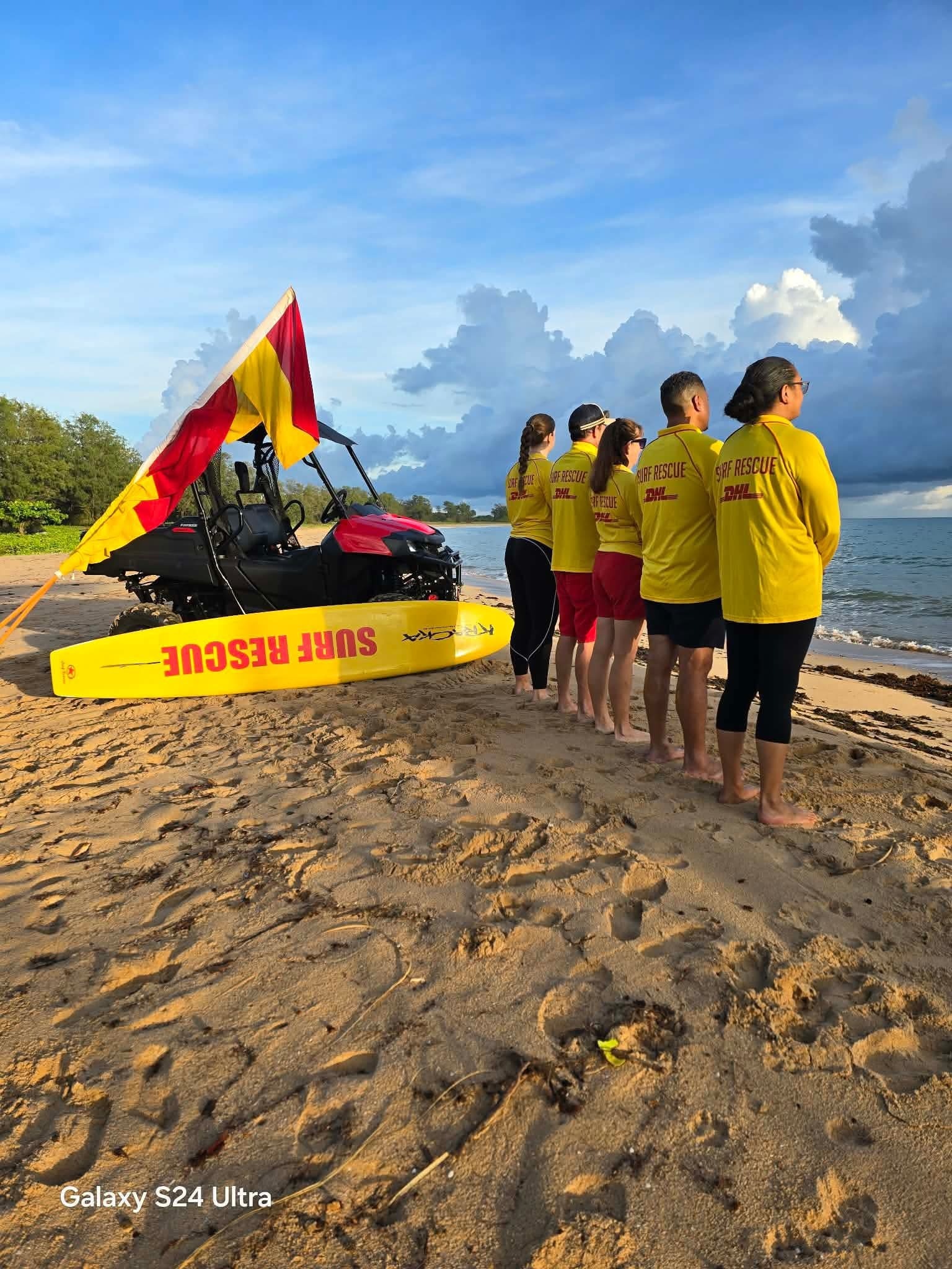 Cinco salva-vidas de surf ficam ao lado de um carrinho de areia e uma prancha de surf na praia, de frente para o mar