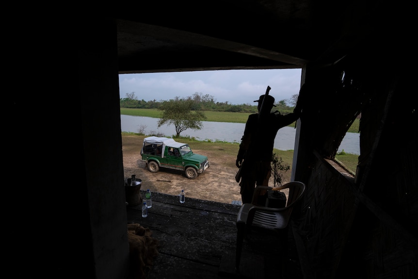 A man stands inside a dark shelter, looking out to an SUV on a grass field, and a body of water.