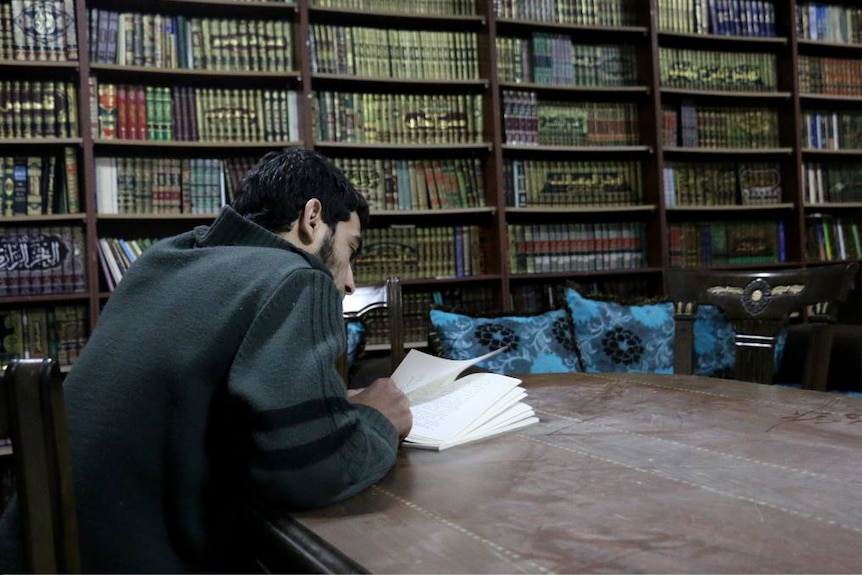 A man sits at a large table reading an open book, surrounded by tall shelves filled with books.