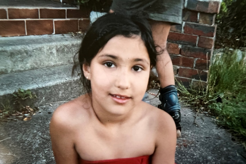 Sabine as a child looks to the camera and smiles as she sits on a concrete path. Yasmin is seen rollerblading behind.
