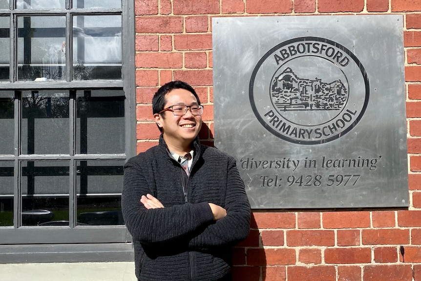 Man in grey jumper with arms crossed smiles in front of silver plaque on brick wall.