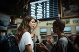 A woman with brown hair points angrily at a board displaying flights as she speaks to another woman.