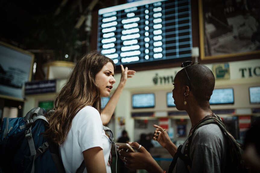 A woman with brown hair points angrily at a board displaying flights as she speaks to another woman.