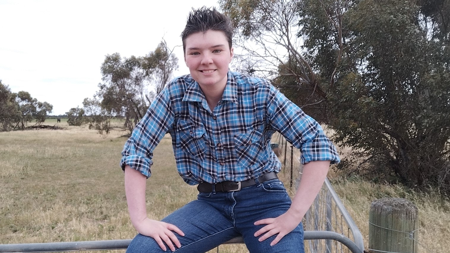 A young man with short brown hair in a check tshirt and jeans, sitting perched on a fence in a paddock. 