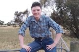 A young man with short brown hair in a check tshirt and jeans, sitting perched on a fence in a paddock. 