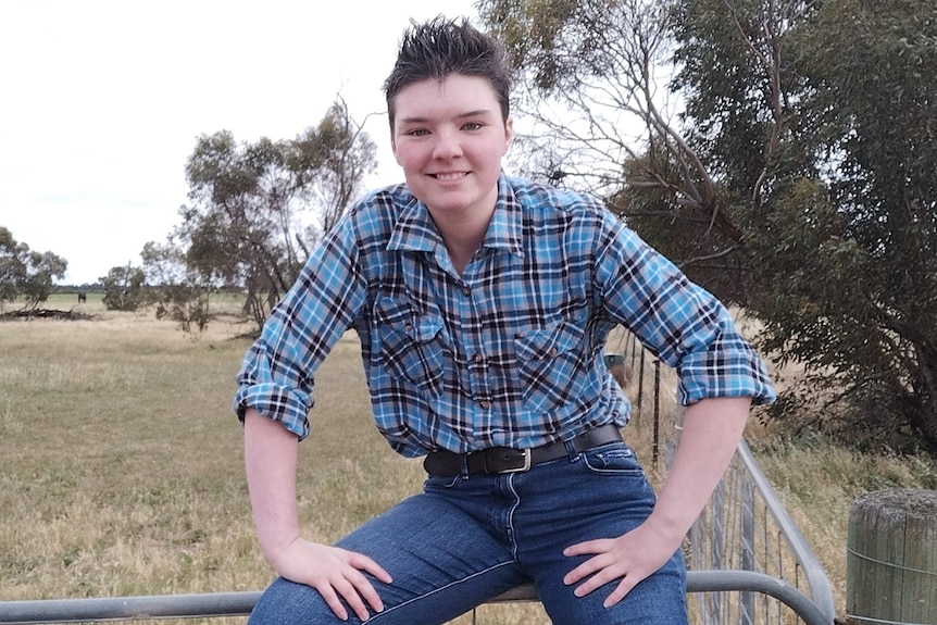 Image of Elliot sitting on a fence, smiling while wearing a blue checkered shirt and dark blue jeans.
