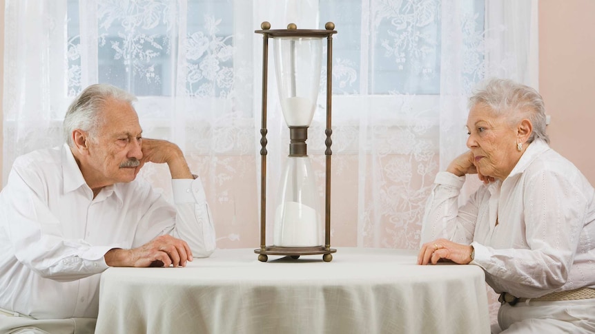 An old man and an old woman sit at a table, looking bored, staring at a giant hour glass half full of sand