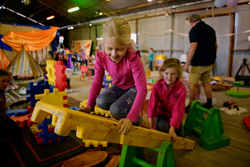 Twee kleine meisjes klimmen op een speelgoedglijbaan in een grote schuur, met overal kinderspeelgoed.