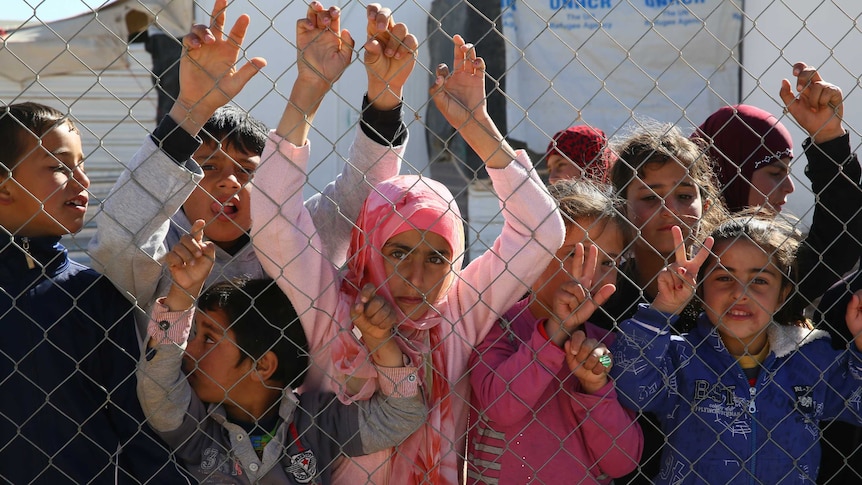 Children stand behind a wire fence staring out from a tented camp.