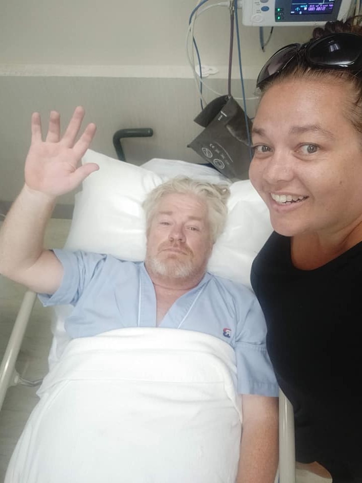 A man in a hospital bed waves alongside his wife who has a big smile