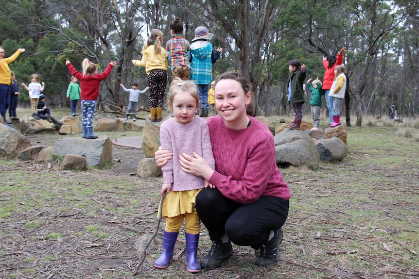 A young mother crouches next to her small child in a bush setting