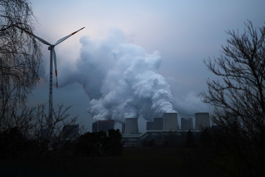 A wind turbine with smoke stacks in the background.