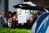 Men holding a white funeral casket covered in flowers next to a car