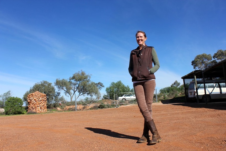 A woman stand on red dirt in front of a shed and a stone water tank.
