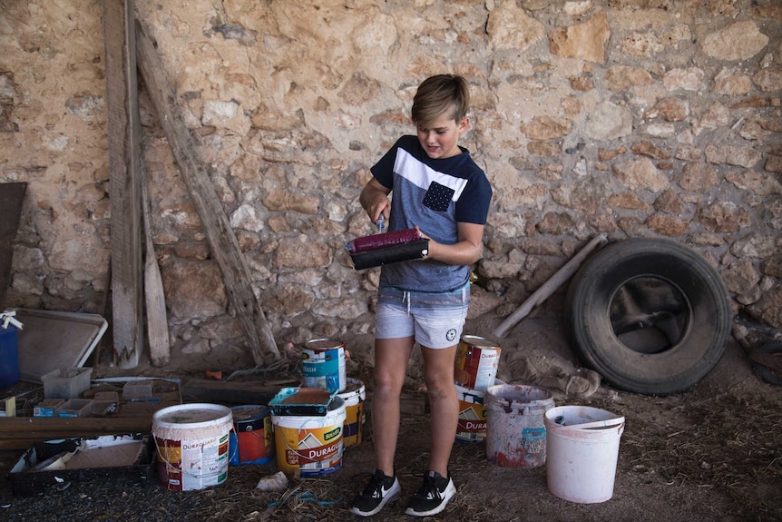 A young boy stands in a shed surround by tins of paint and holding a paint pallet.