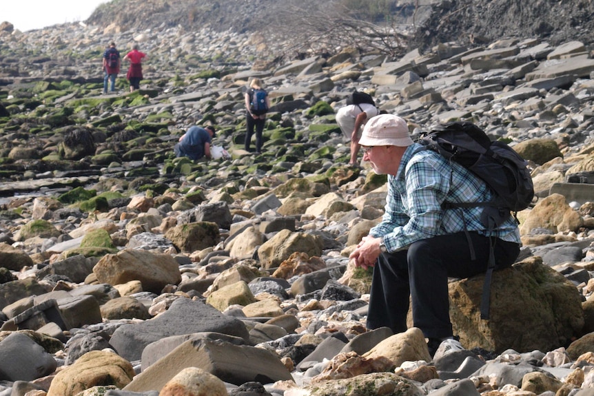 An elderly man sits on a rock with a backpack on while others search the rocky coastal landscape.