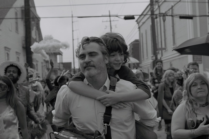 Black and white image of a middle aged man with a beard wearing a white shirt and giving young boy a piggyback ride in a crowded street