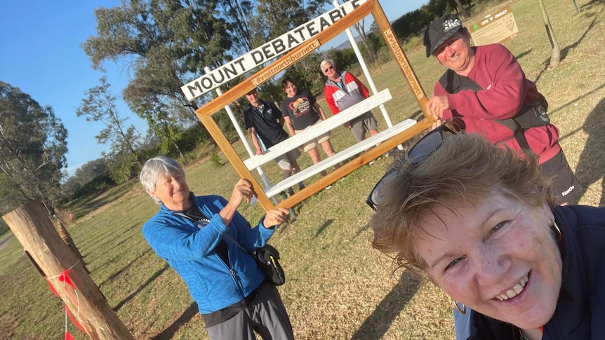 A woman in the front of camera with people behind holding a sign that says "Mt Debatable."