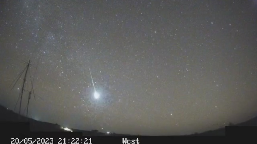 A bright meteor falls from a starry sky. The image is captured on a grainy black and white camera. 