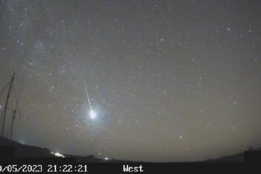 A bright meteor falls from a starry sky. The image is captured on a grainy black and white camera. 