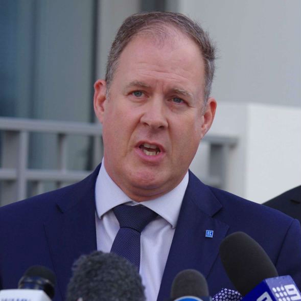 man in suit talking, with microphones in front of him