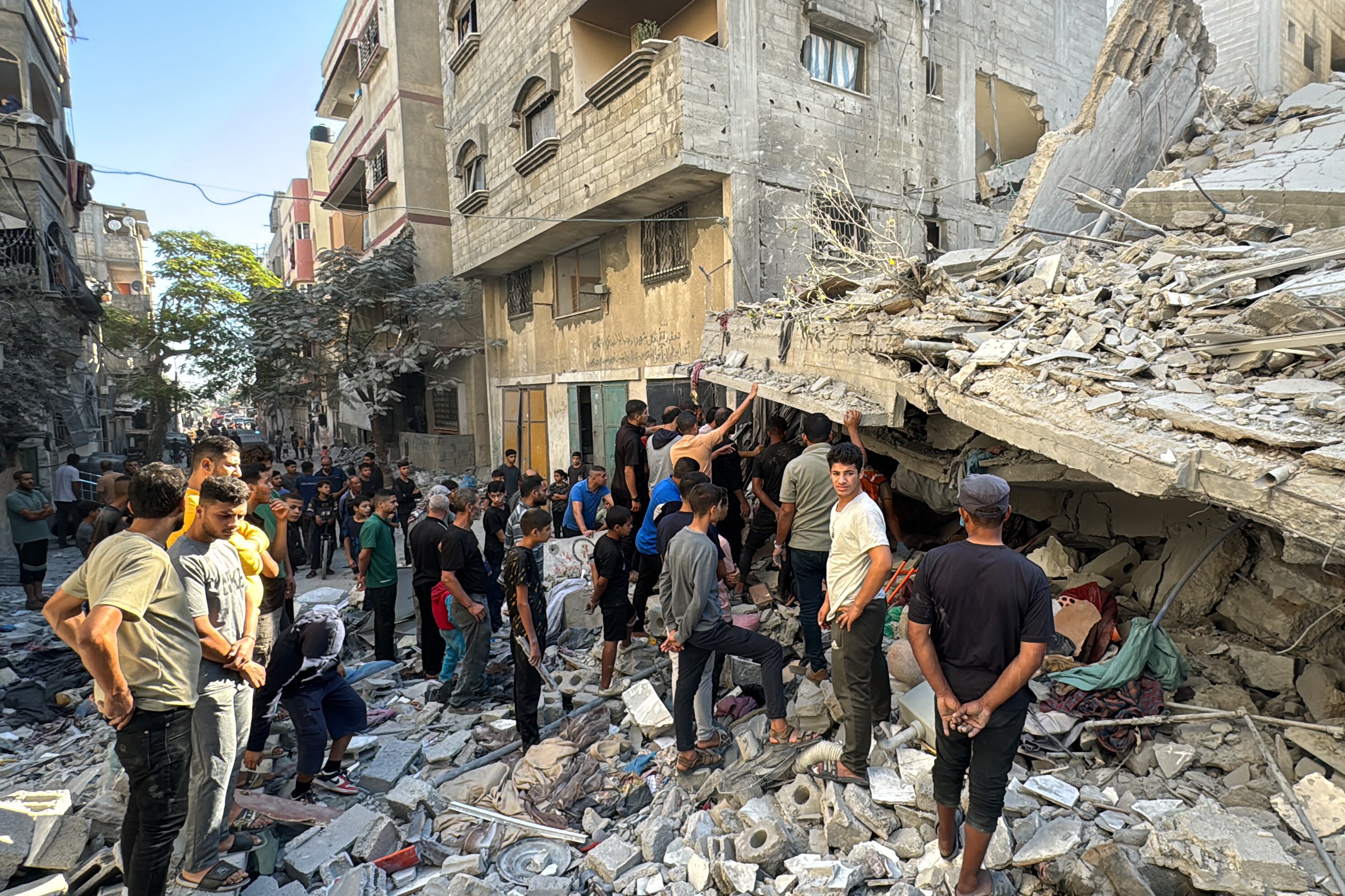 A crowd gathers outside damaged buildings, with one in the foreground reduced to rubble.