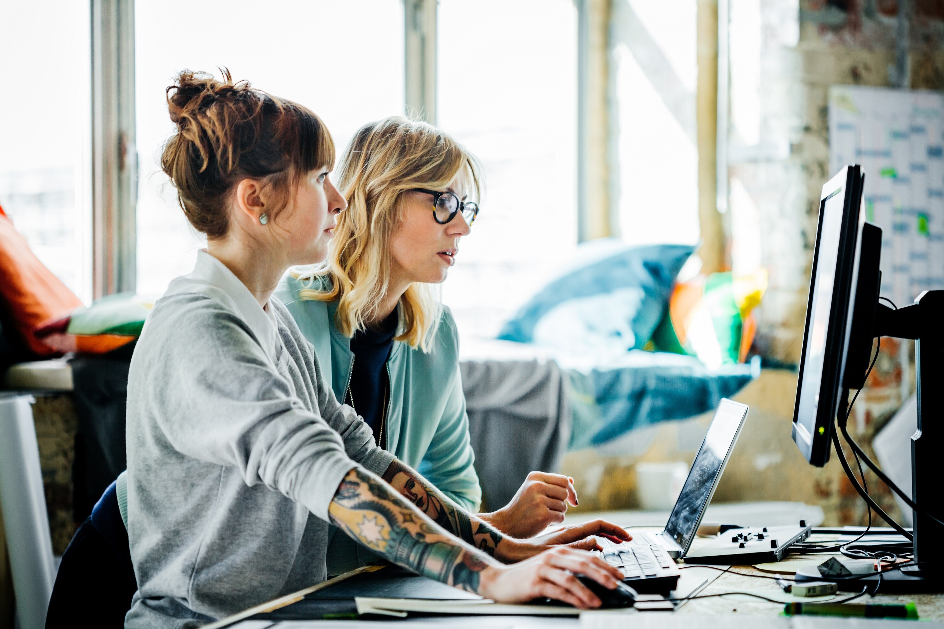 Two young women, one with red hair and tattoos, the other blonde with glasses, look at a computer screen