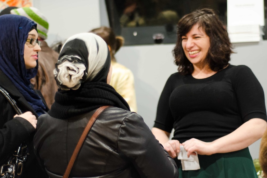 A woman with dark hair speaks to two women wearing headscarves at an event opening.