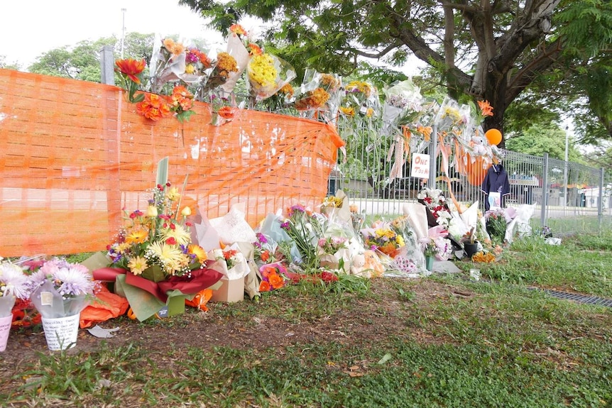 Flower tributes line a fence line with orange mesh. .