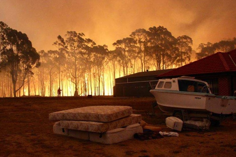 Orange flames and smoke light up bush, a house and boat are in the foreground