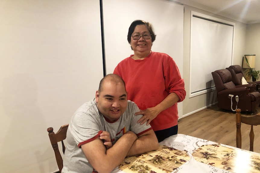 A woman with short brown hair, glasses and a red jumper smiles and puts her hands on her son's shoulders as he sits at the table