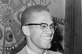 Malcolm X wearing a suit and speaking, in a black and white photo.