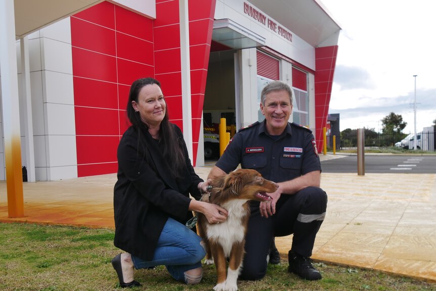 A man and a woman kneel down with a brown dog in front of a fire station building.
