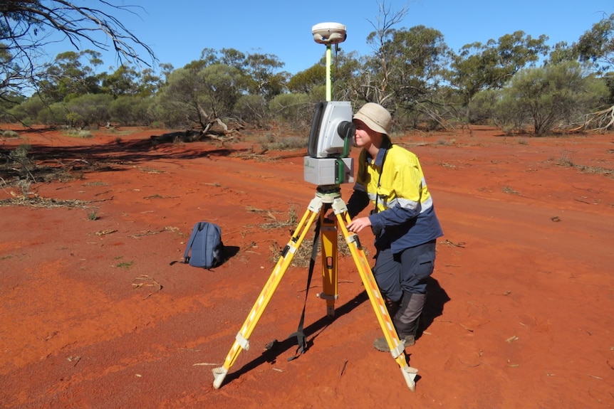 A woman looking into a Lidar machine on a tripod, on the red dirt of the Midwest.