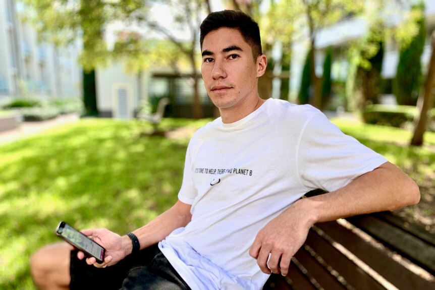 A young man wearing a white T-Shirt, sitting on a park bench holding a smart phone.