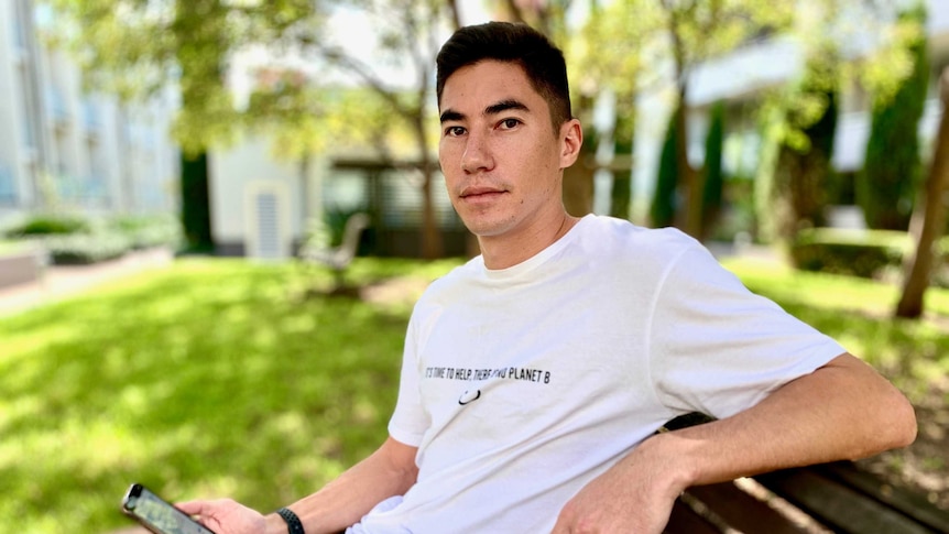 A young man wearing a white T-Shirt, sitting on a park bench holding a smart phone.