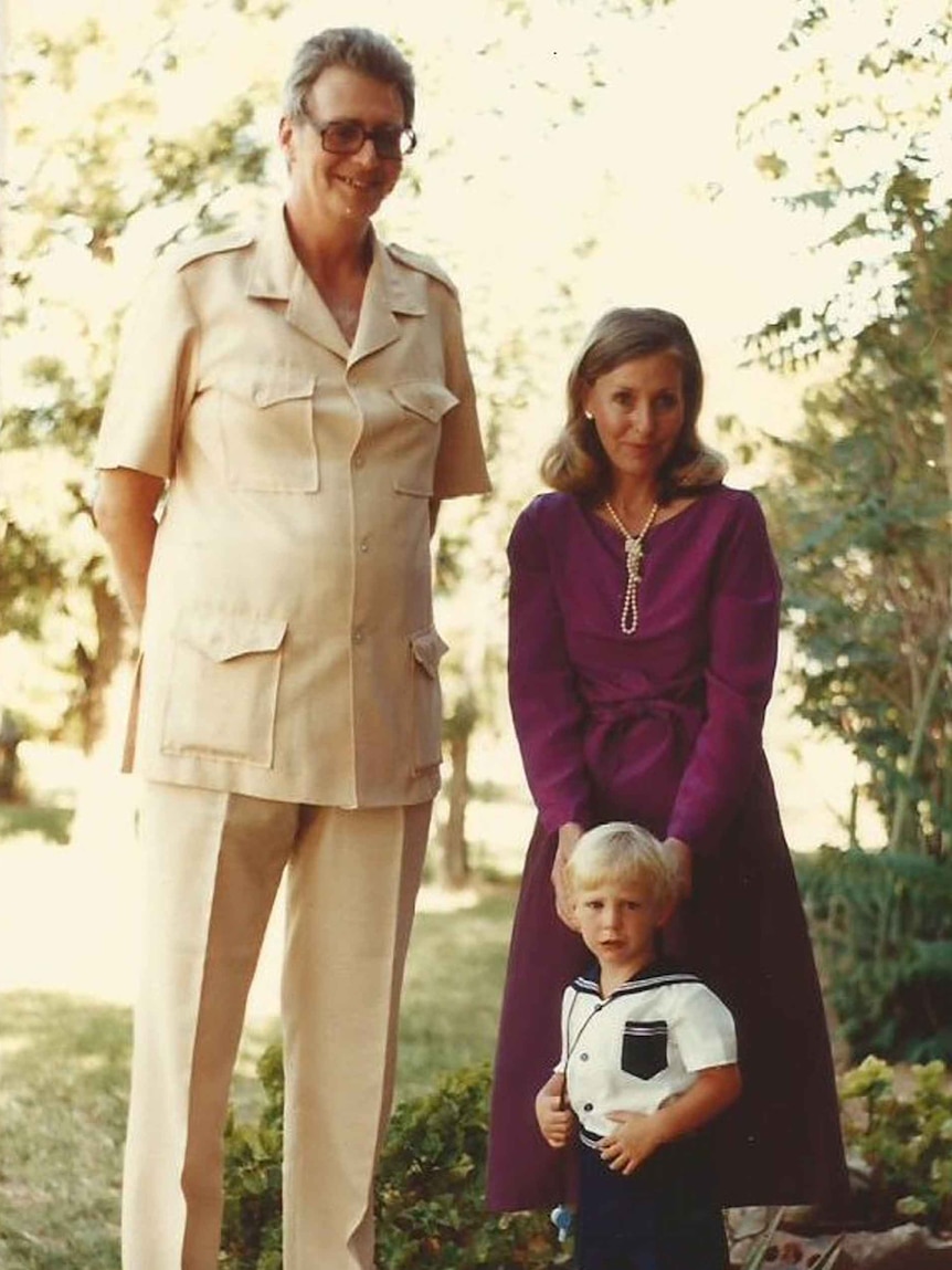 A family portrait of a tall man in safari suit, petite woman in purple dress, and child in sailor's costume
