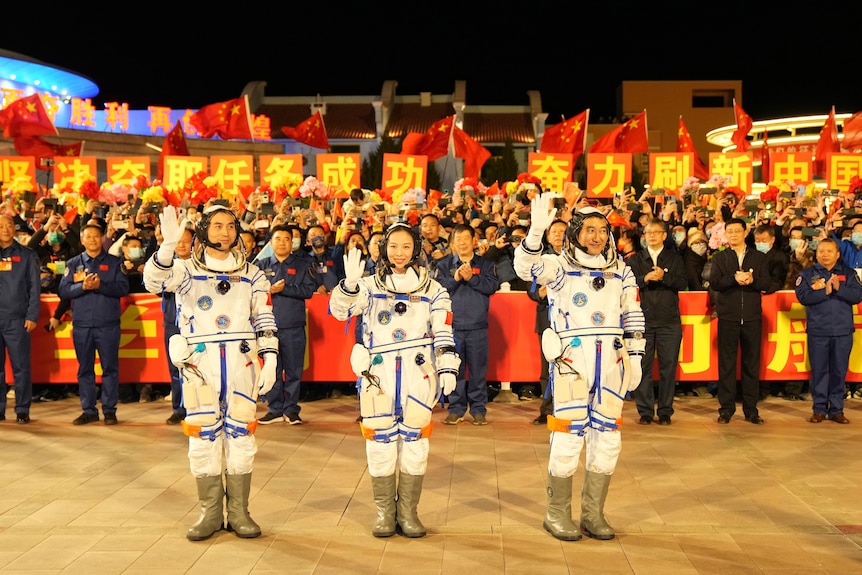 Three Chinese astronauts stand and wave in front of a crowd.