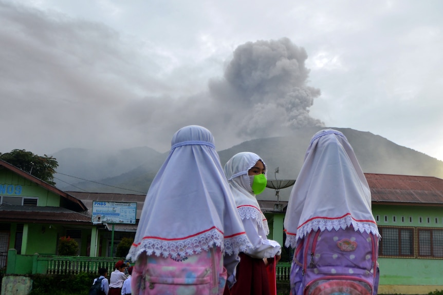 Three female students in colouful clothes and white headscarves look from their schoolyard towards an erupting volcano.