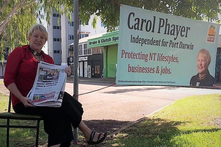 Woman sitting on a chair in a park holding up a newspaper beside an election banner.