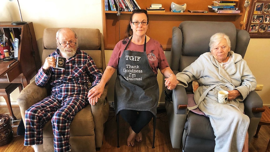 An old man and an old woman sit in armchairs while a woman sits between them, holding their hands.