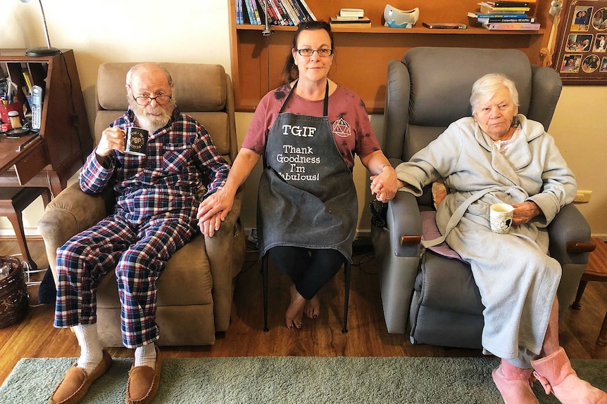 An old man and an old woman sit in armchairs while a woman sits between them, holding their hands.