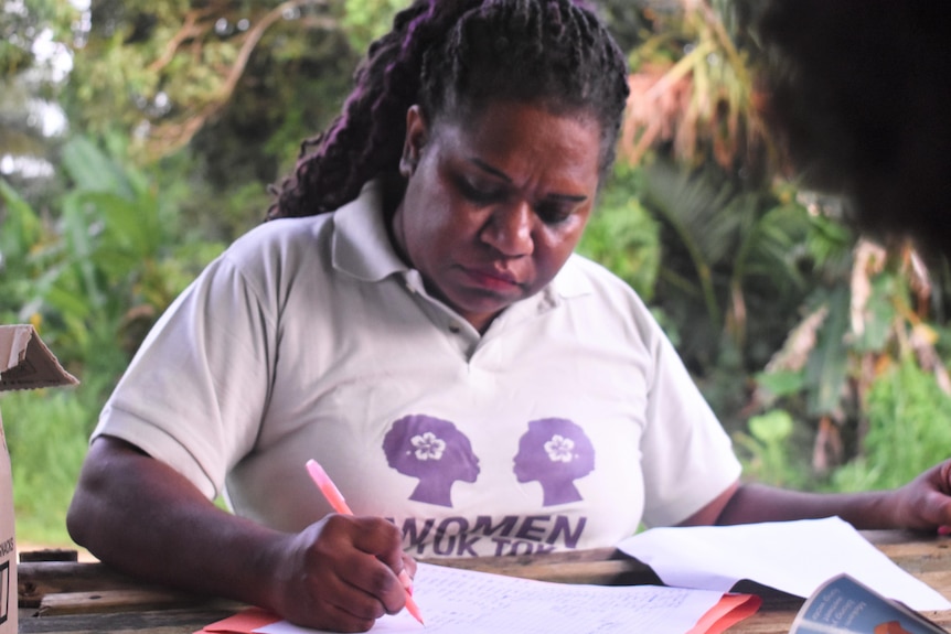 Ni-Vanuatu woman Flora Vano sitting down writing in a book wearing a white and purple t shirt