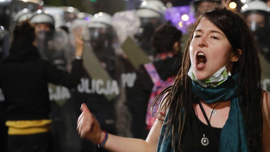 A female protester with dreadlocks and a mask around her chin yells, standing in front of a line of police with riot shields.