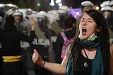 A female protester with dreadlocks and a mask around her chin yells, standing in front of a line of police with riot shields.
