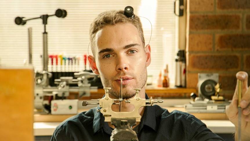 A young man sits in a workshop surrounded by fine, hand-crafted tools.