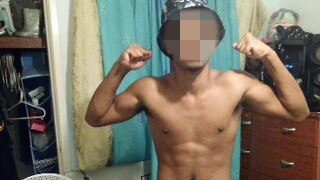 A boy stands with his biceps flexed in what looks like his bedroom, with his shirt off.