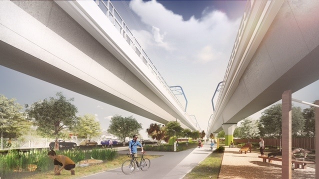 A man walks his bike below two parallel raised train tracks, with green, public spaces either side of him.